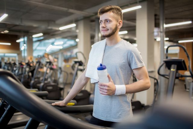 Sporty guy with towel and bottle of water leaning on one of treadmills before or after sports training